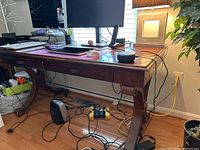 Angled front and side view of the wooden desk showing the decorative curved legs, two front drawers with metal pulls, the metal bar across the base, and some wiring underneath.