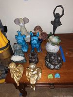 Full view of all figurines on a wooden surface showing two blue ceramic dogs, multiple frogs, angels, and a black ballerina figurine.