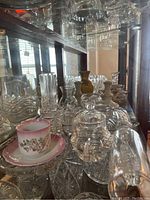 Multiple clear glass and crystal serving and decorative items in cabinet including covered dishes, vases, and a tea cup set.