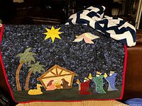 Photo showing a handmade nativity scene quilt alongside a large blue and white geometric patterned blanket on a couch.