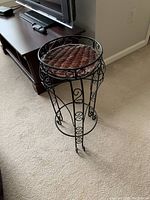 Full side view of the black iron plant stand with woven rattan shelf placed on carpet, showing the curved legs and scroll detail.