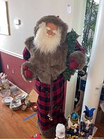 Full body photo of a life size Santa figure wearing a red plaid robe and a brown fur vest, holding greenery with string lights wrapped on the arm.