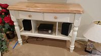 Wood table with natural wood top, two drawers with wooden knobs, open lower shelf, and distressed off-white painted base with turned legs. Shown in living room setting near lamp and speakers.