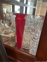 Tall red glass vase surrounded by clear cut glass items in cabinet