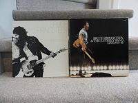 Front covers of both LPs placed on carpeted stairs: 'Born to Run' and 'Live 1975-85' box set with Bruce Springsteen playing guitar in black and white and stage light photo respectively.