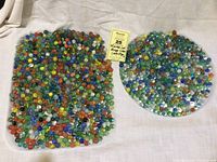 Two large piles of assorted vintage glass marbles with colorful swirls and cats eyes, displayed on a white cloth with a label showing lot number and description.