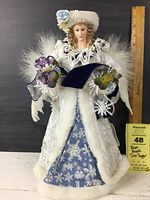 Full frontal view of angel Christmas tree topper wearing blue velvet gown with snowflake designs, white faux fur trim, lace collar, and feather wings. Purple floral accents held in hands. Ruler in background shows height approx. 16.5 inches.