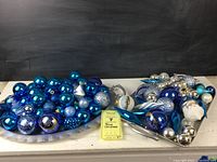 Overall lot showing blue and silver ornaments arranged on two silver trays, including round baubles, teardrop shaped ornaments, and Santa head figure.