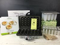 Lot showing the waffle sticks maker open, with two sets of pop molds and muffin rings behind it, plus wooden sticks in a small bag.