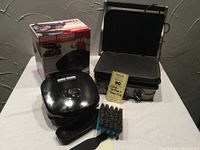 Photo showing George Foreman Healthy Eating Grill in front of its box, Cuisinart Panini and Grill Maker with open grill plates, metal scrub brush and MaxSold tag.