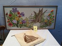 Full view of both vintage crewel wall hangings displayed on a white table with a blue wall background. The larger piece shows a floral forest scene and the smaller a sailboat.