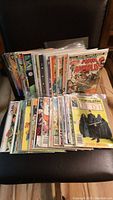 Photo shows a large stack of vintage comic books with colorful covers featuring various characters. Comics are in plastic sleeves and stacked upright and flat.