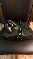 Front view of original Xbox console with controller and cables placed on top, showing green Xbox logo and controller details
