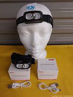 Photo showing two black LED headlamps with elastic head straps, two white USB charging cables, and two white packaging boxes labeled 'LED HEADLAMP'. One headlamp is displayed on a mannequin head.