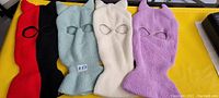 Four different colored knit ski masks with knit horns, displayed side by side on a yellow surface, with eye holes and knit horn details.