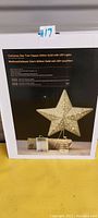 Photo of the product packaging showing details of the gold glitter Christmas star tree topper with LED lights and spring base.