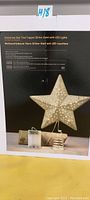 Boxed gold glitter star Christmas tree topper with LED lights and gold spring base visible on the packaging.