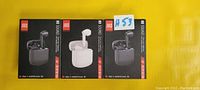 Three boxed J80 wireless earbuds with charging cases, two black and one white, displayed on a yellow surface.