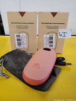 Two boxes of rechargeable handwarmers with one pink handwarmer, USB charger, wrist strap, and pouch visible on yellow surface.