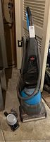 Full upright view of black and blue Bissell carpet cleaner with hose and attachments in basement setting.