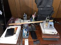 Overview of the lot showing cordless phone, wooden ruler, binders, multiple pens in holders, and calculators on a wooden desk against paneled wall.
