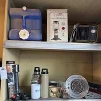 Lot photos showing the boxed Coleman propane lantern, blue water jug, Airthereal heater, thermoses, camping stove, cups, and fishing cutters on shelves.