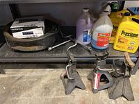 Photo showing two metal car jack stands, oil drain pan, assorted automotive fluid containers including Prestone antifreeze, and the boxed Kenwood car stereo on a garage shelf.