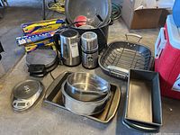 Overview of kitchenware lot showing various pans, baking trays, kitchen scale, kettle, grinder, waffle iron, and mixing bowls on floor.