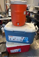 Orange thermos jug with white lid and spigot on top of a blue and white Coleman cooler with label showing features and capacity, stacked above red and white cooler, in garage environment.