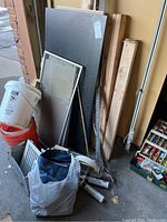 Image shows stacked drywall sheets and various pieces of wood lumber standing upright against a wall, plastic buckets, and other construction materials piled on ground.