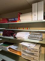 Shelves with board games, white binders, packing tape, printer paper, hole punch, and plastic drawers organizer under shelf.