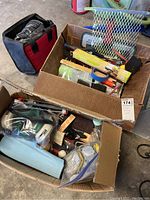 Two cardboard boxes filled with various painting supplies including a paint sprayer, brushes, rollers, sponges, and gloves.
