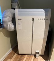 Front view of Sharp air conditioner with flexible exhaust hose attached and remote control placed at base.