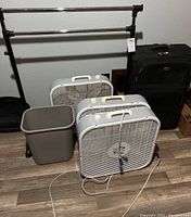 Photo of three white box fans, black suitcase, gray waste bin, and adjustable black metal clothing rack. Items arranged on wooden floor against white wall.