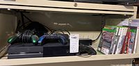 Xbox 360 console with 4 controllers on a shelf alongside cables and original box in the background