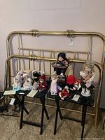 Wide view showing nine porcelain dolls of various sizes seated and standing on black stools against a brass bed frame background