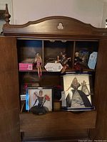 Wood cabinet with Barbie dolls seated and boxed inside, some loose dolls visible along with boxed dolls.