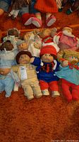 Ten dolls arranged on carpet, showing varied clothing and styles including hats and pacifiers.