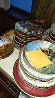 Stack of collector plates showing varying designs and sizes, some with colorful gold and pink rims, and a plate featuring outdoor scene with a person and dog, needs cleaning.