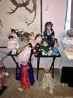 Photo showing multiple porcelain collector dolls arranged on tables and chairs posed in front of a floral wall hanging, displaying various sizes and colorful detailed period outfits.