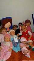 Group photo of eight dolls including large Rosie O'Donnell dressed doll seated on the floor among several baby and toddler dolls in various outfits.