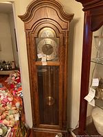 Full view of the tall wooden Howard Miller grandfather clock with glass cabinet door and carved top