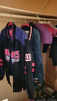 Black NFL Giants jacket with purple and pink accents and logo visible, hanging on rack.