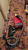 Full view of red canister vacuum with ribbed hose, extended metal wand, main suction floor head and additional brush attachment laid out on floral carpeted floor