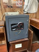 Front and side view of gray Sentry metal safe on wooden stand with combination lock handle and red-accented dial.