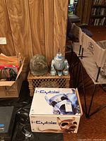 Full lot showing the I-Cybie box, silver robot figurine and camouflage helmet on a wicker crate against a wood-paneled wall.