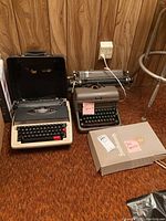 Both typewriters side by side on brown floor against wood panel wall, electric typewriter in case on left, Royal manual typewriter on right with paper and box of Supplement brand typewriter paper.