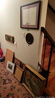Photo showing a variety of wall art pieces including framed drawings, cork board and round decorative plate leaning against a wall next to a staircase.