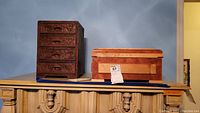 Two wood storage boxes on a pedestal; one carved with 4 drawers and floral patterns, the other rectangular with top lid and smooth finish.