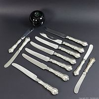 Photo showing 7 sterling silver knives and 4 butter spreaders arranged flat with a green apple for size reference.